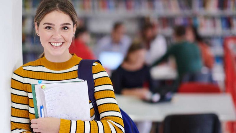 a portrait of a girl holding a notebook in her hand while standing in front of the library